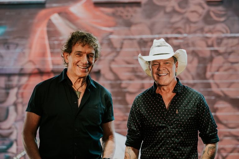 Australian musicians Ian Moss and Troy Cassar-Daley smiling together outdoors in front of a mural, wearing casual shirts and a cowboy hat.